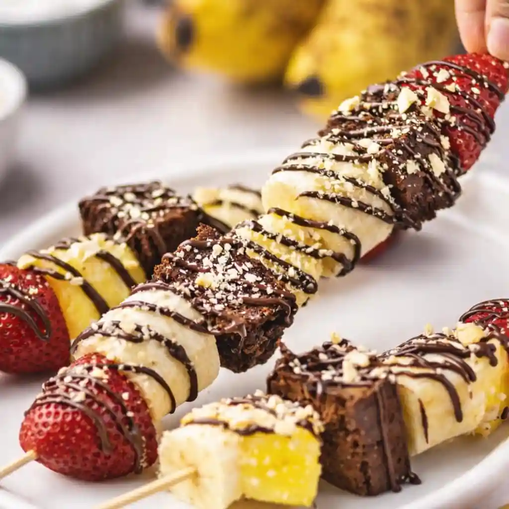 Banana split dessert kabobs with chocolate drizzle on white marble background