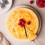 A wide porcelain stand holding a traditional vibrant crème brûlée cheesecake on a bright white marble background.